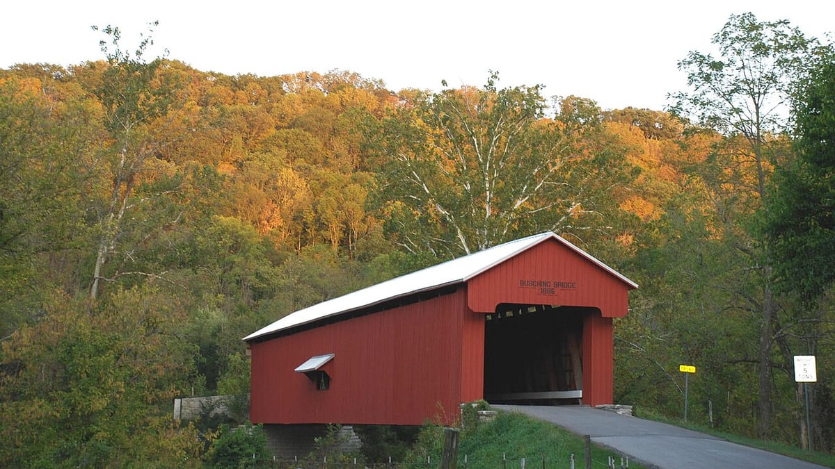 A red covered bridge on a country road below a hillside turning gold in early autumn